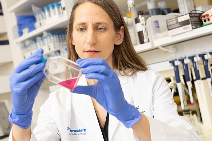 Woman with long brown hair in a lab, wearing a lab coat and blue gloves, looking at a red substance in a beaker.