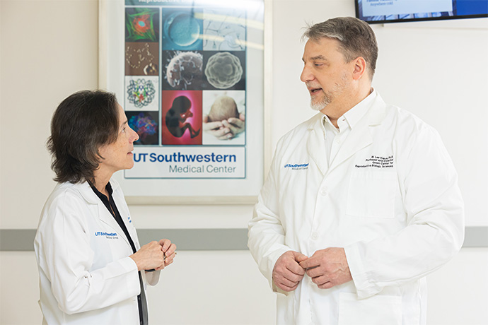 A man and woman wearing lab coats talking in a hallway in front of a UT Southwestern Mecial Center poster.