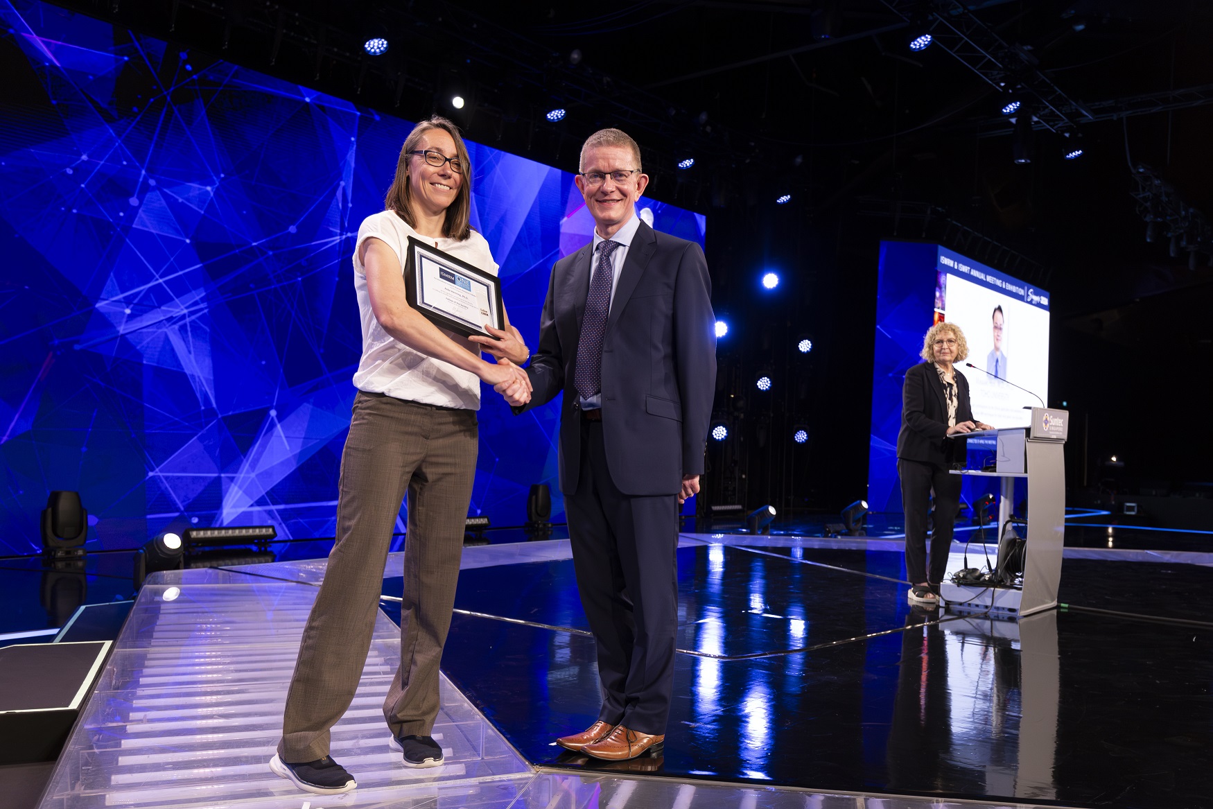 Smiling woman with brown hair and dark-rimmed glasses, holds a placque and shakes hands with a smiling man with short brown hair and glasses, while blond woman stands at a podium, watching. 