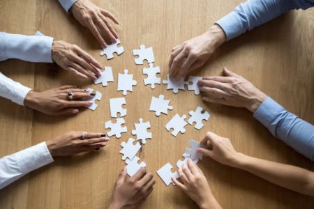 9 hands reach in a circle around white puzzle pieces on a table