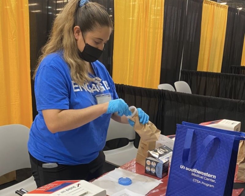 woman in blue tshirt with long ponytail and black medical mask and surgical gloves works at event table