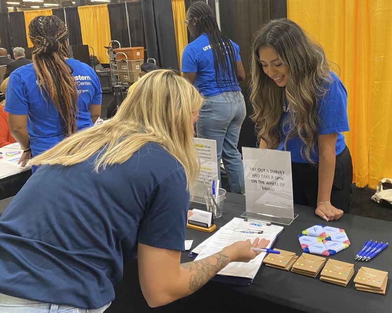 smiling woman with long hair assists blond woman filling out form at event table; 2 other female UTSW workers in background