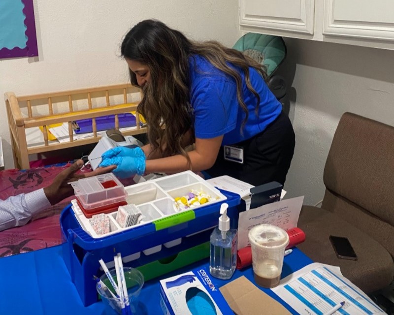 UTSW medical working with long dark hair wearing surgical gloves does finger stick blood test on hand of patient seated at table with medical supplies