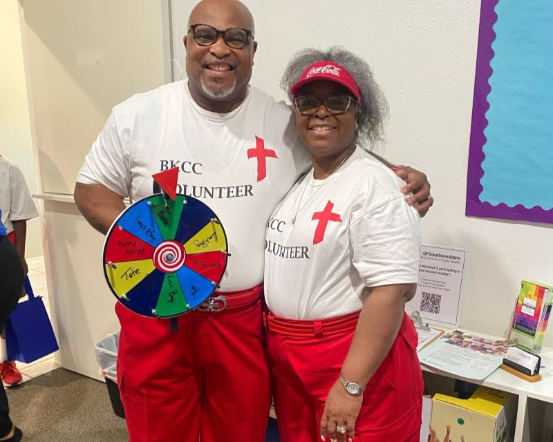 2 smiling older black people wearing volunteer tshirts and red pants spin prize wheel at event