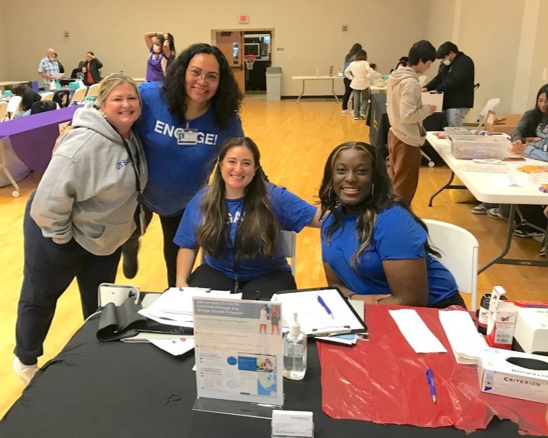 large room with 2 rows of tables in background, front table manned by 4 UTSW community engagment workers: woman with blonde hair standing, woman with black hair standing, woman with long dark hair seated, black woman iwth black hair seated