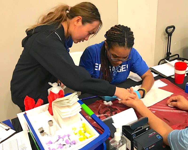 young medical worker with ponytail holds patient's hand as another UTSW medical woker in ENGAGE! tshirt performs stick test on finger