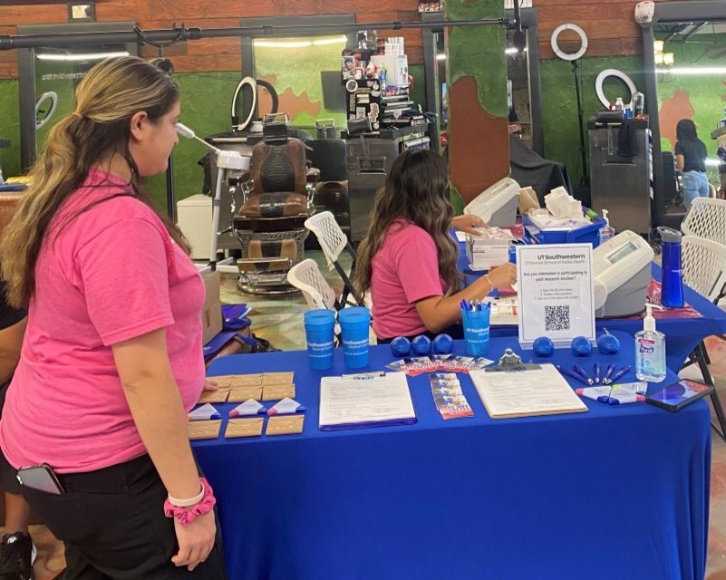 2 women with pink tshirts and long dark hair work 2 event tables with UTSW swag during community barber shop event