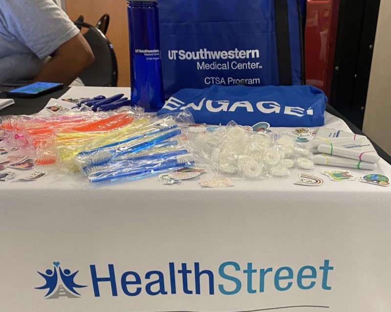 UT Southwestern and Healthstreet logos on tabletop display of freebies such as keyrings, stickers, tote bags and tshirts