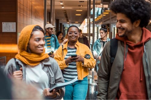 group of 6 young diverse students walk down a hallway toward camera; left front girl wears yellow head scarf, right front man in grey jacket over red hoodie, black woman in center wears glasses, yellow jacket and striped shirt