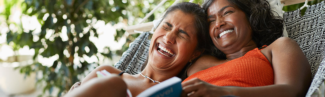 2 laughing women recline on outdoor hammock with book on their laps