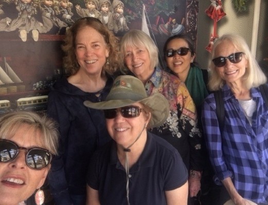 in front of wall painting of cherubs in hooded cloaks, group of smiling women - 2 without sunglasses, 4 with sunglasses