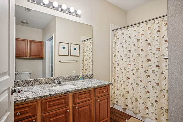 Brown bathroom cabinets, sink with granite-like counter, larger mirror, shower curtain pulled across shower