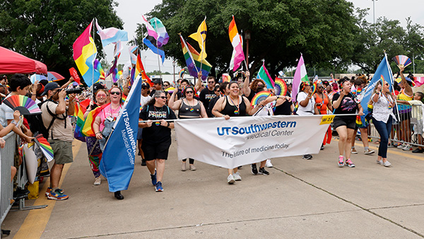 A group of men and women in summer attire parade down a street carrying multi-color flags and banners that read - UT Southwestern Medical Center, The Future of Medicine