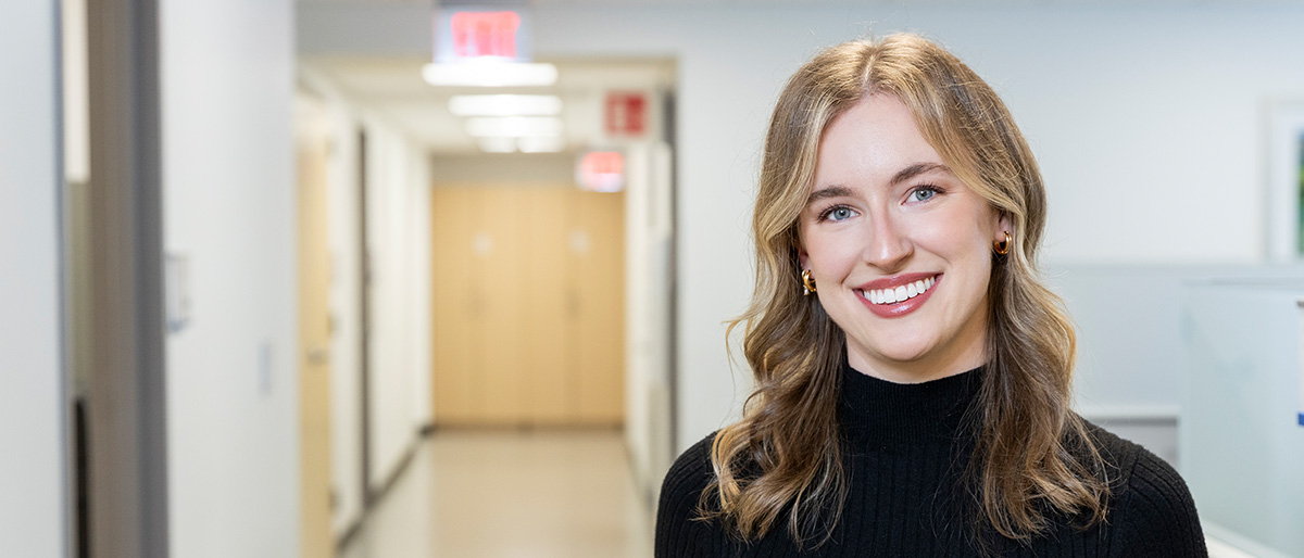 Smiling woman with long blond hair, wearing a black turtleneck and gold earrings, standing at the end of a long hallway.