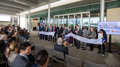 12 people holding up a ribbon in front of the doors of the new TI BMES building, while a man in the center cuts the ribbon. A seated audience watches.