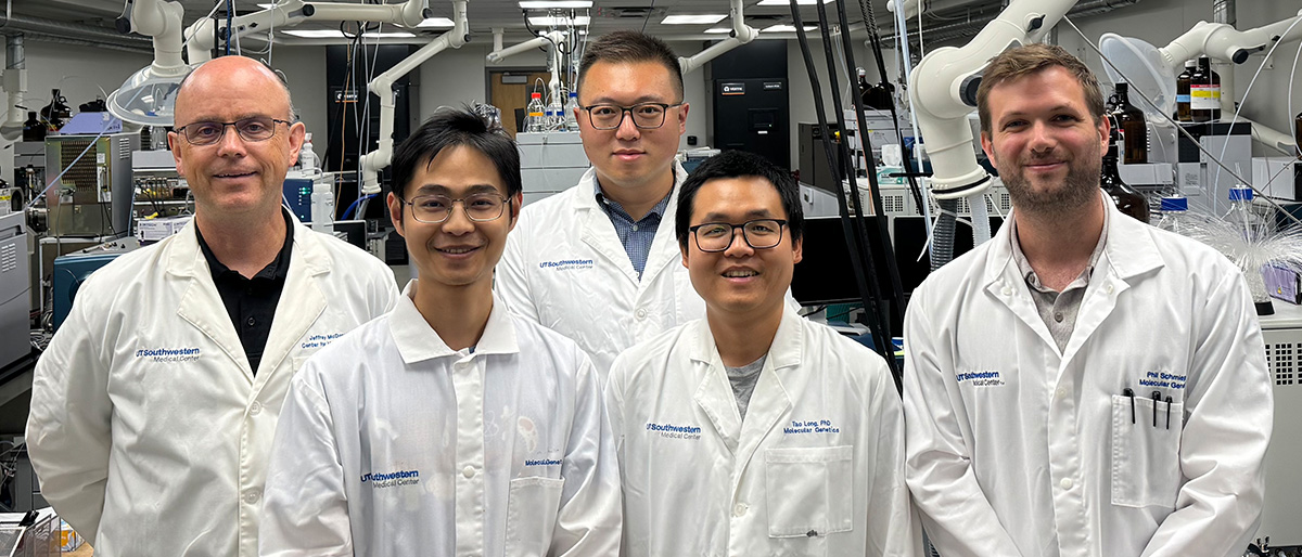 5 men wearing white UT Southwestern lab coats standing in a lab in front of large scientific equipment.