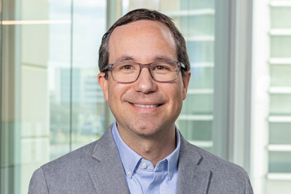 Smiling man with brown hair and dark-rimmed glasses, wearing gray suit, gray shirt, and a blue tie.