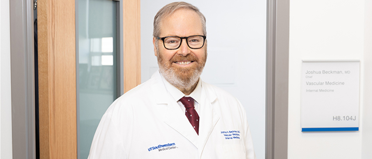 Smiling man with graying hair, beard, and mustache, wearing a white UT Southwestern lab coat and dark rimmed glasses.