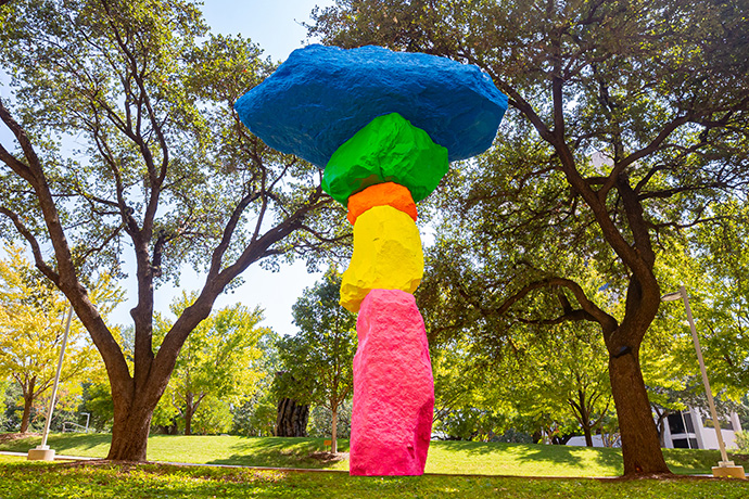 Tall outdoor sculpture shaped vaguely like a tree built large bright boulders, painted pink, yellow, green, and blue.