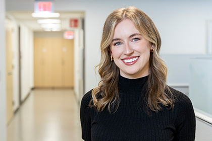 Smiling woman with long blond hair, wearing a black turtleneck and gold earrings, standing at the end of a long hallway.