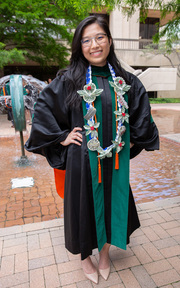 New graduate Dr. Tran Nguyen of Estabrook College poses with her regalia and a special homemade necklace with flowers crafted from dollar bills. She is all smiles about staying at UT Southwestern for her residency.