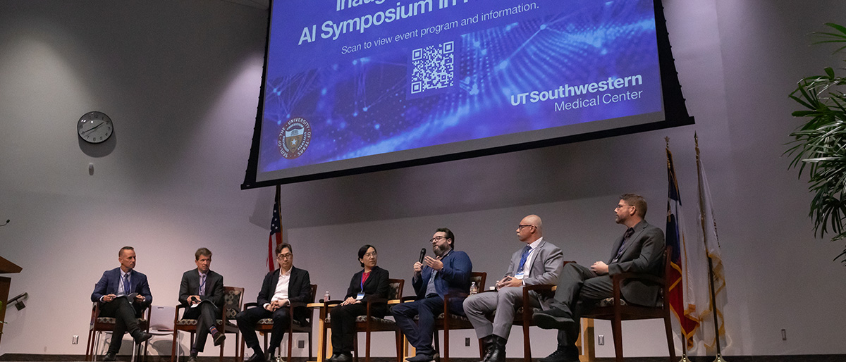 6 individuals dressed in suits, seated on a stage, with a screen behind them announcing the Inaugural UT System AI Symposium in Health Care at UT Southwestern Medical Center.