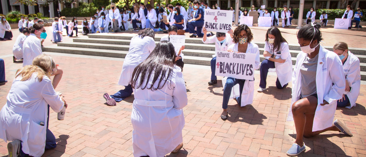 People in white lab coats holding protest signs, kneeling in a courtyard