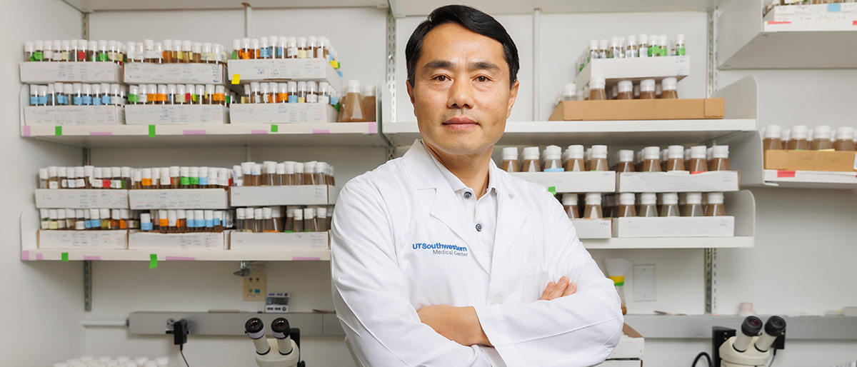 Dr. Pan, a man with dark hair wearing a lab coat, standing in front of shelves in the lab.