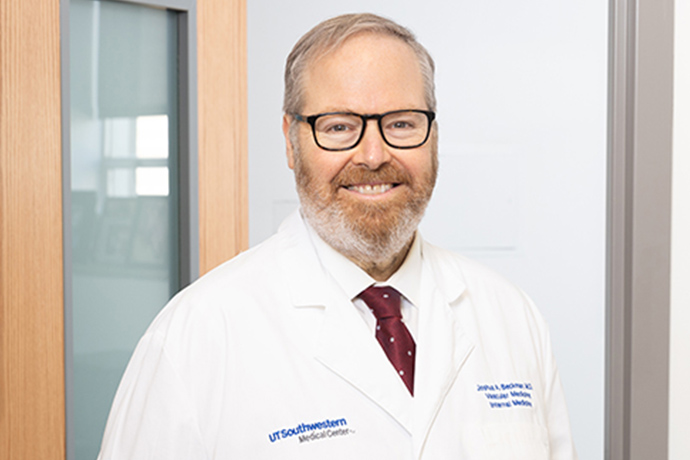 Smiling man with graying hair, beard, and mustache, wearing a white UT Southwestern lab coat and dark rimmed glasses.