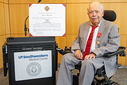Smiling man with gray hair wearing a gray suit, white shirt, and red tie, with a red and silver medal pinned to his lapel. Next to him on a UT Southwestern Medical Center podium is a large certificate.