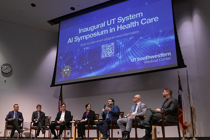 6 individuals dressed in suits, seated on a stage, with a screen behind them announcing the Inaugural UT System AI Symposium in Health Care at UT Southwestern Medical Center.
