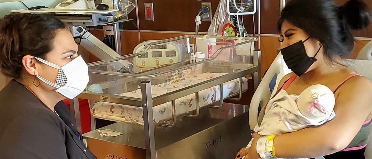 Two womenin masks next to hospital baby bed, one is holding a child