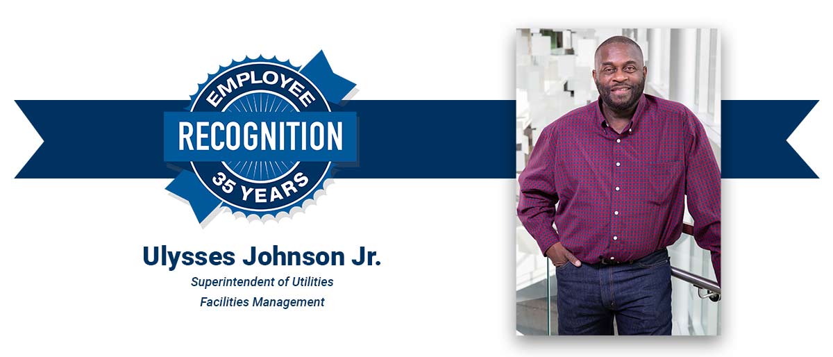 Smiling man with cropped dark hair, beard, and mustache, wearing a magenta checked shirt and jeans. On white banner with-Ulysses Johnson Jr., Superintendent of Utilities, Facilities Management, and blue Employee Recognition Program logo.