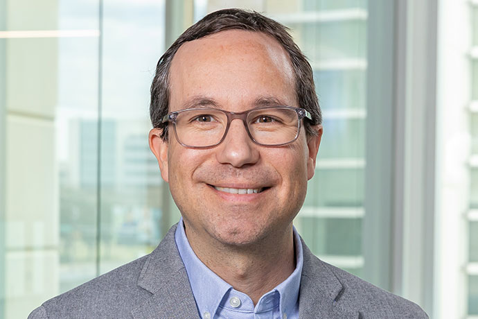 Smiling man with brown hair and dark-rimmed glasses, wearing gray suit, gray shirt, and a blue tie.