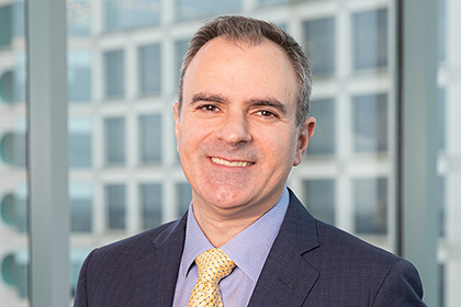 Smiling man with receeding gray hair, wearing a dark gray suit, blue shirt, and yellow tie.