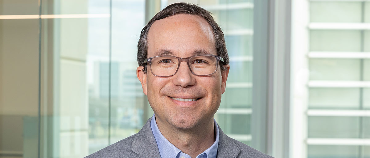 Smiling man with brown hair and dark-rimmed glasses, wearing gray suit, gray shirt, and a blue tie.