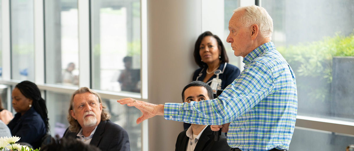 Standing man with white hair wearing a green, blue, and white plaid shirt, speaking to a group of men and women, some seated at a table, on which is a jar of white flowers.