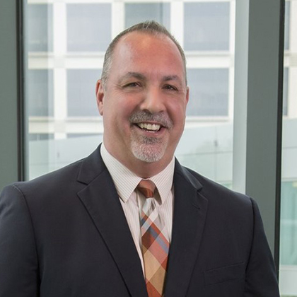 Smiling man with fair receding hair wearing a dark suit, light shirt, and brown plaid tie.