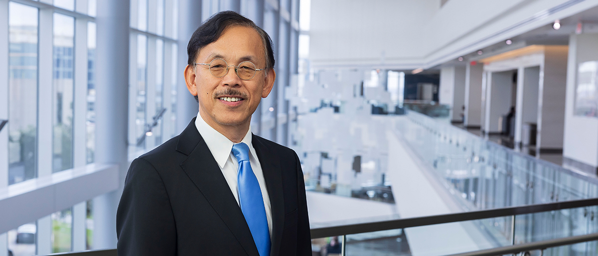 Smiling man dark graying hair and mustache, wearing a dark suit, white shirt, and blue tie.