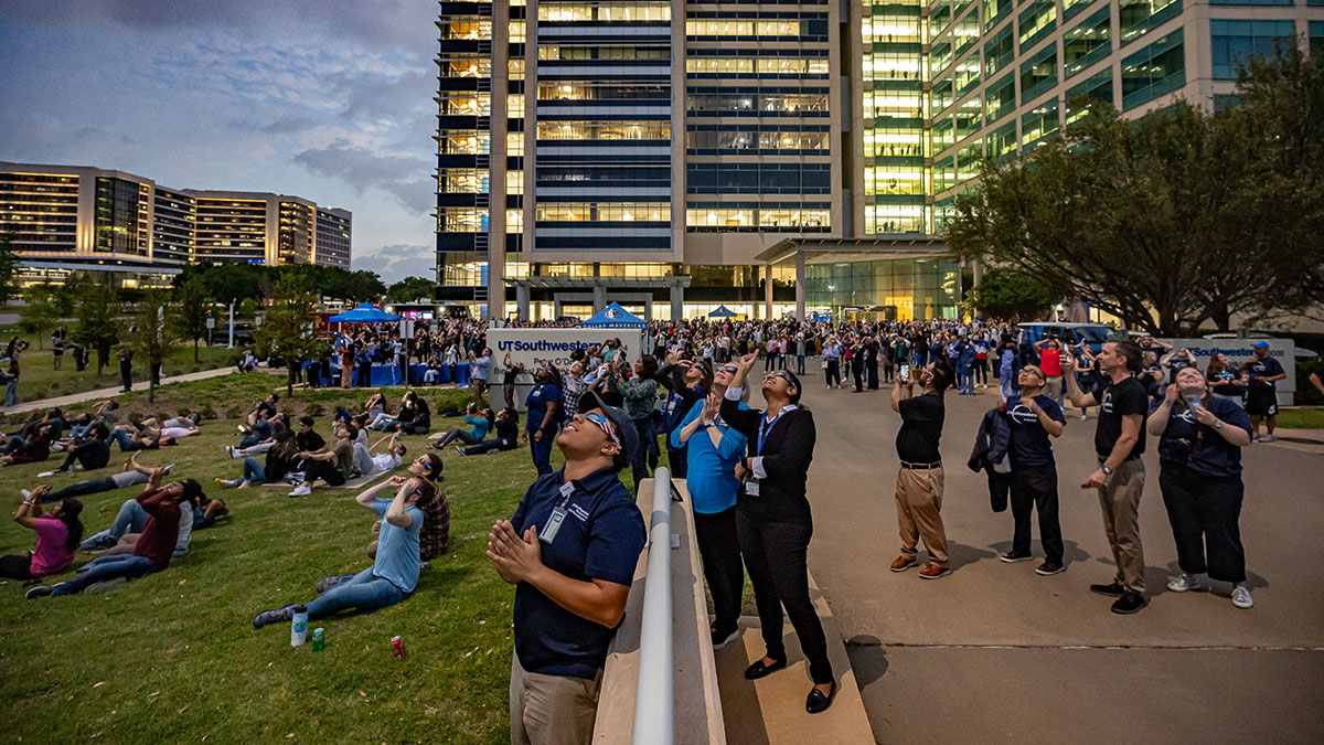 A group of people stand and sit on the ground looking at the darkened sky outside UT Southwestern