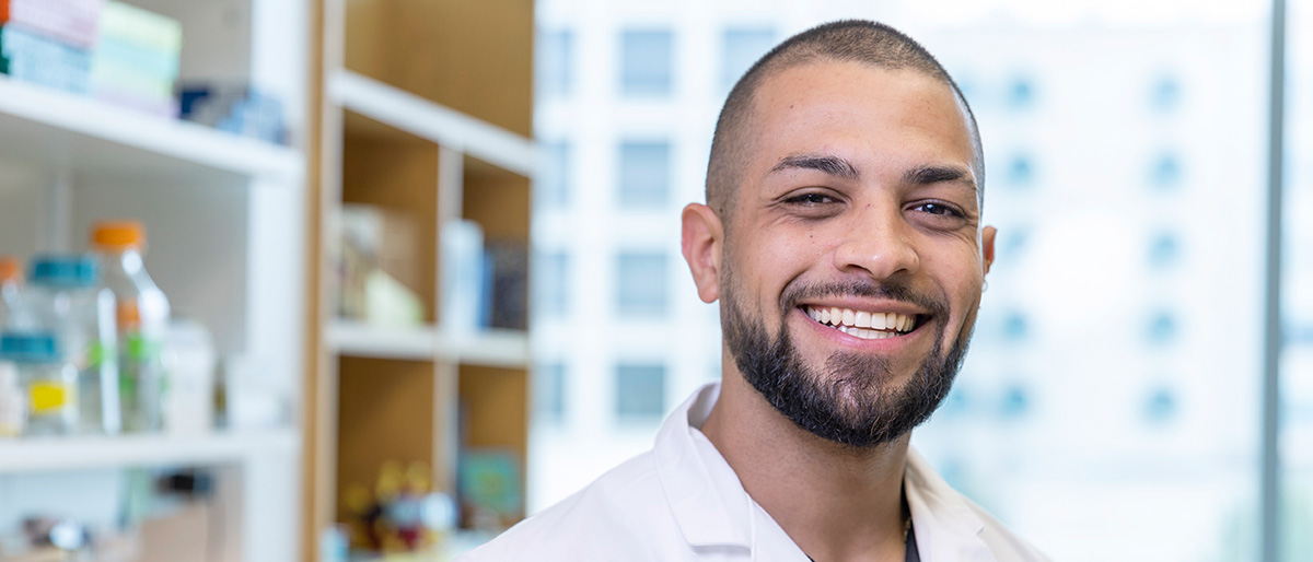 Smiling man with trim dark hair, beard, and mustache, wearing a white UT Southwestern Medical Center lab coat.