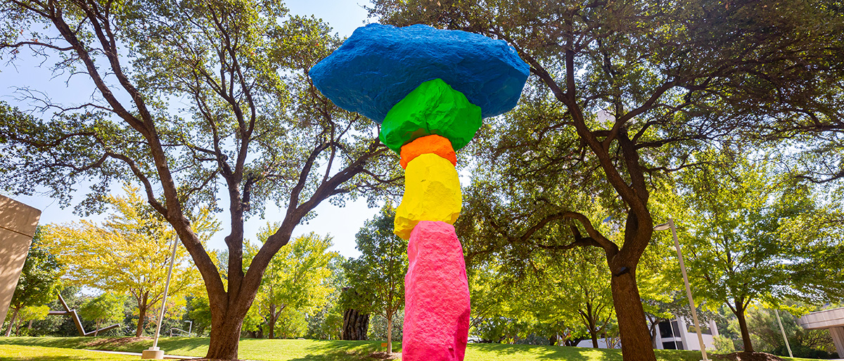 Tall outdoor sculpture shaped vaguely like a tree built large bright boulders, painted pink, yellow, green, and blue.