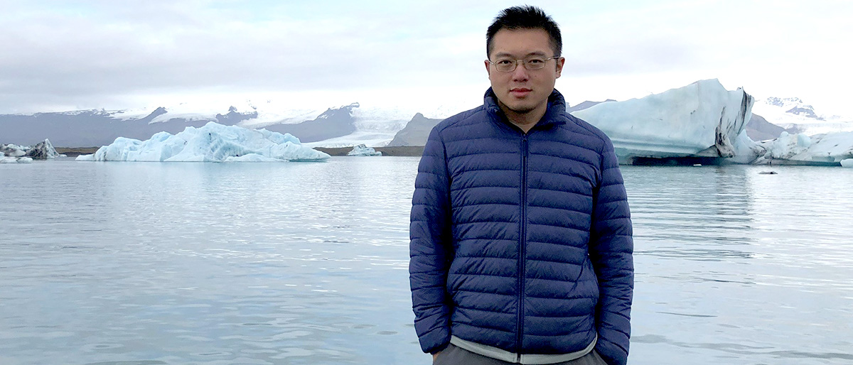 Dark-haired man wearing glasses and a blue puffer jacket standing in front of glacier and ice flow.