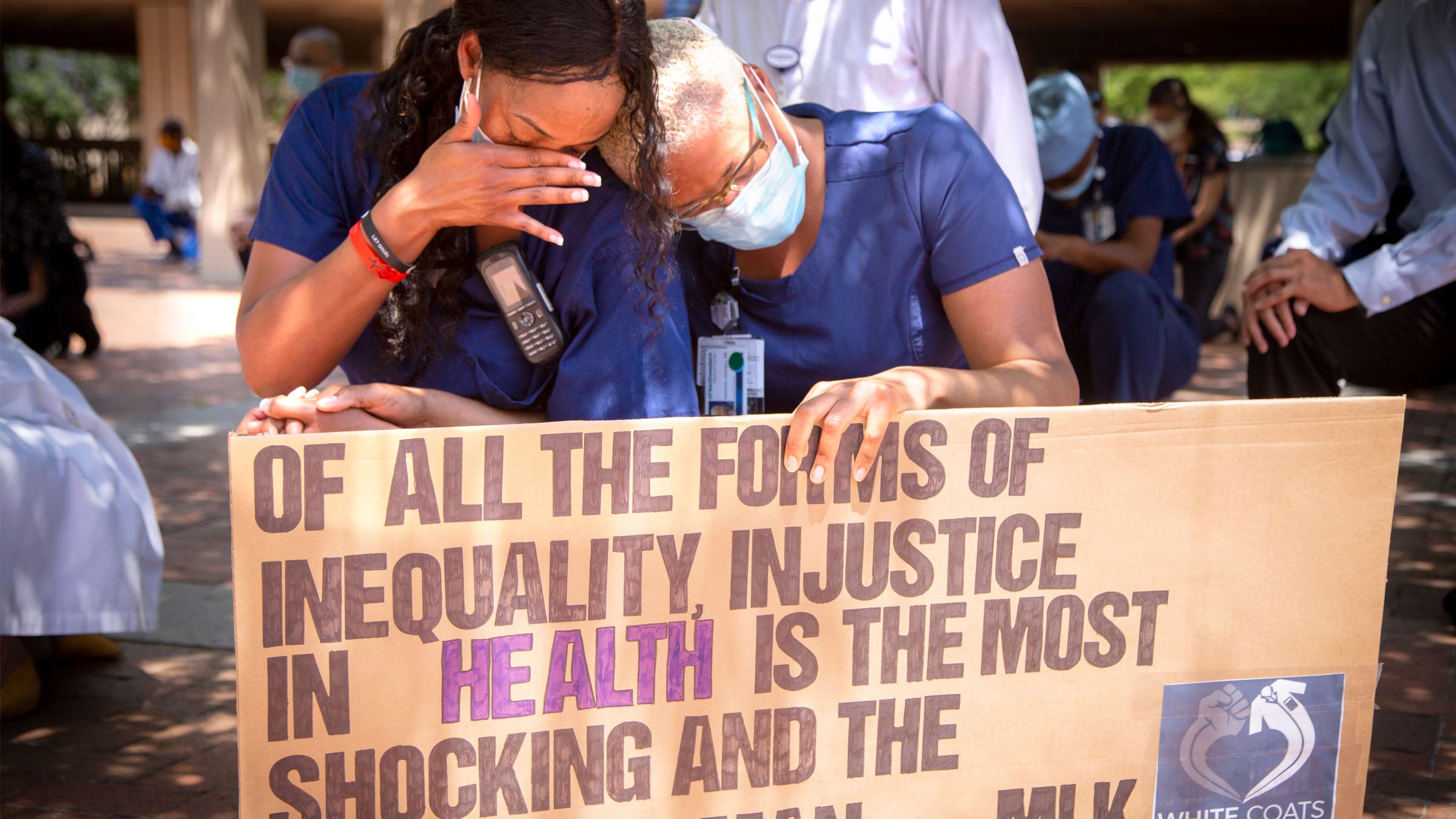 Two women in scrubs and masks, holding a sign that reads Of all the forms of inequality, injustice in health care is the most shocking and inhumane