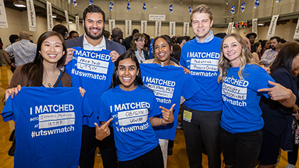 Six smiling students hold up blue t-shirts with their match information printed on it