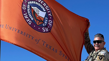 grey haired man in sunglasses and military camo uniform holds orange University of Texas System flag