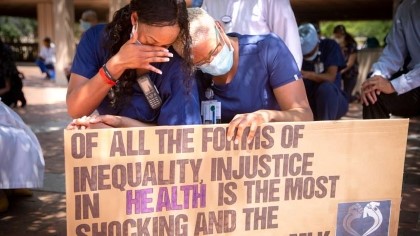 Two women in scrubs and masks, holding a sign that reads Of all the forms of inequality, injustice in health care is the most shocking and inhumane