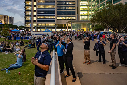 exterior lawn of hospital where crowds of people wearing solar eclipse glasses look up at the sky