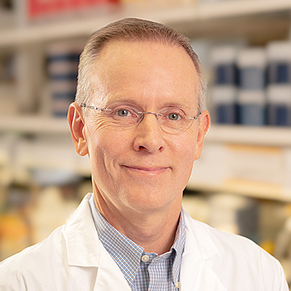 smiling man with thinning gray hair, wire-rimmed glasses wearing white lab coat standing in from of shelves with books and medical equipment