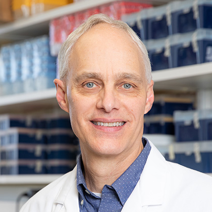 smiling man with thinning white hair, white lab coat and blue collared shirt standing in front of shelves full of medical supplies